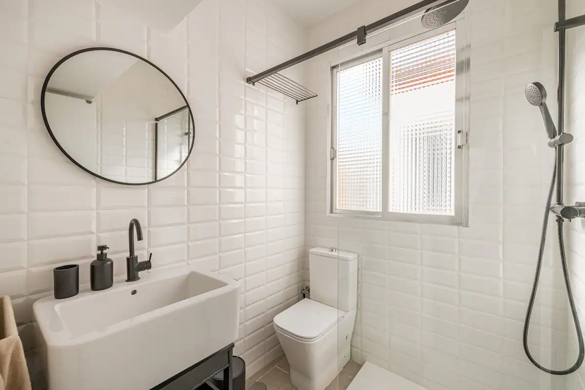 White-tiled bathroom with a round mirror, walk-in shower and a window